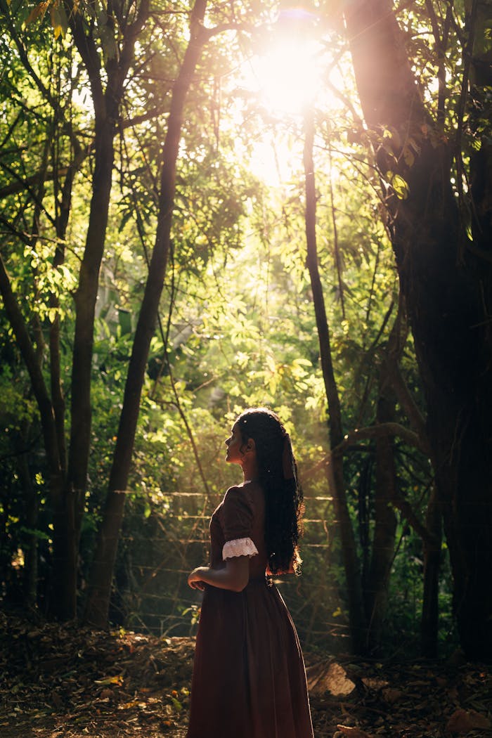 A woman in a dress stands gracefully in a sunlit forest, embodying tranquility.